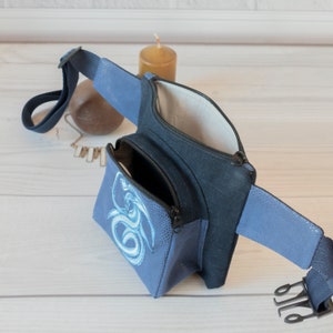 May include: A blue fanny pack with a zippered pocket and a smaller zippered pocket on the front. The front pocket has a blue and white embroidered design of a dragon.
