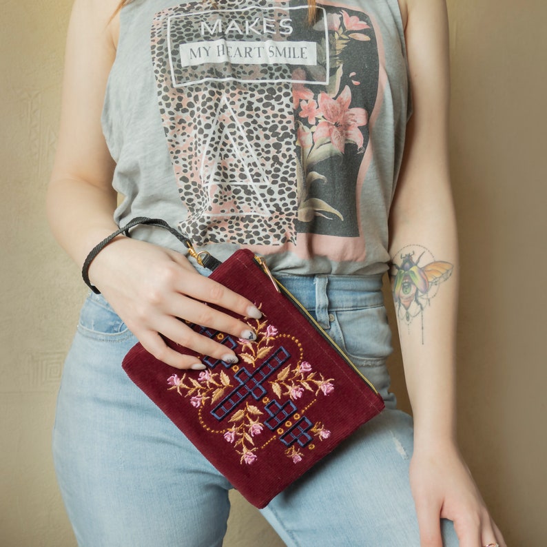 May include: A woman is holding a burgundy velvet clutch with a gold zipper and embroidered floral design. The clutch has a black strap and is worn over the shoulder.