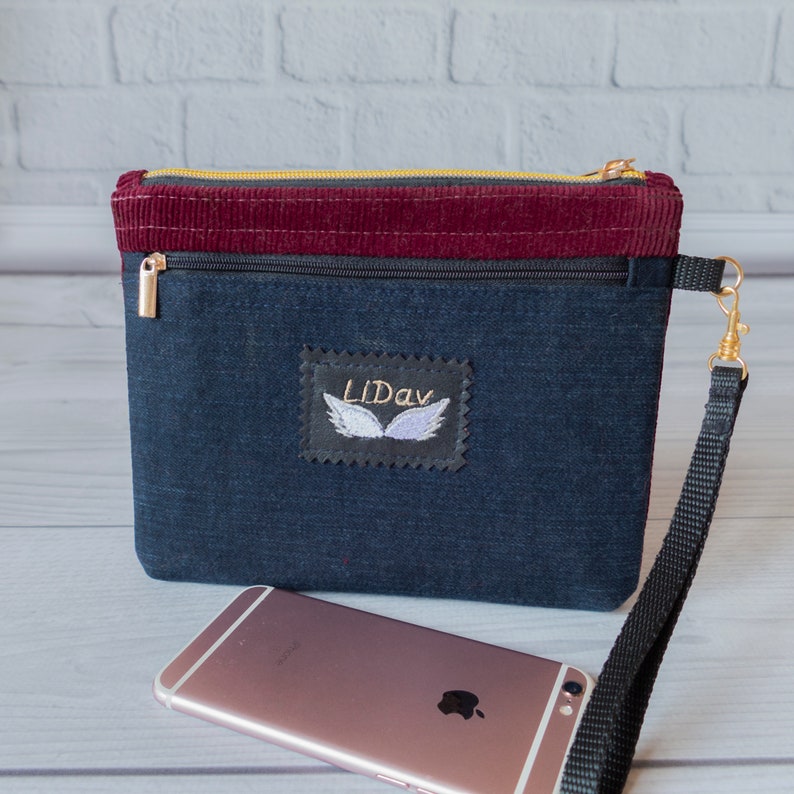 May include: A dark blue denim zippered pouch with a burgundy corduroy top. The pouch has a black strap and a small embroidered patch with the text "LiDav" and a pair of wings.