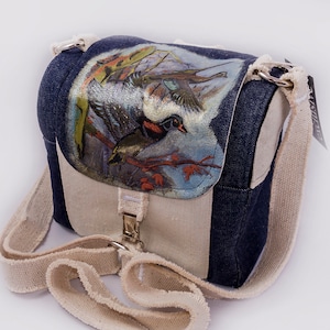 Puede incluir: Un pequeño bolso bandolera de lona azul y blanco con una solapa que presenta una imagen pintada de un pato y otras aves en una rama. El bolso tiene una correa blanca y una hebilla plateada.