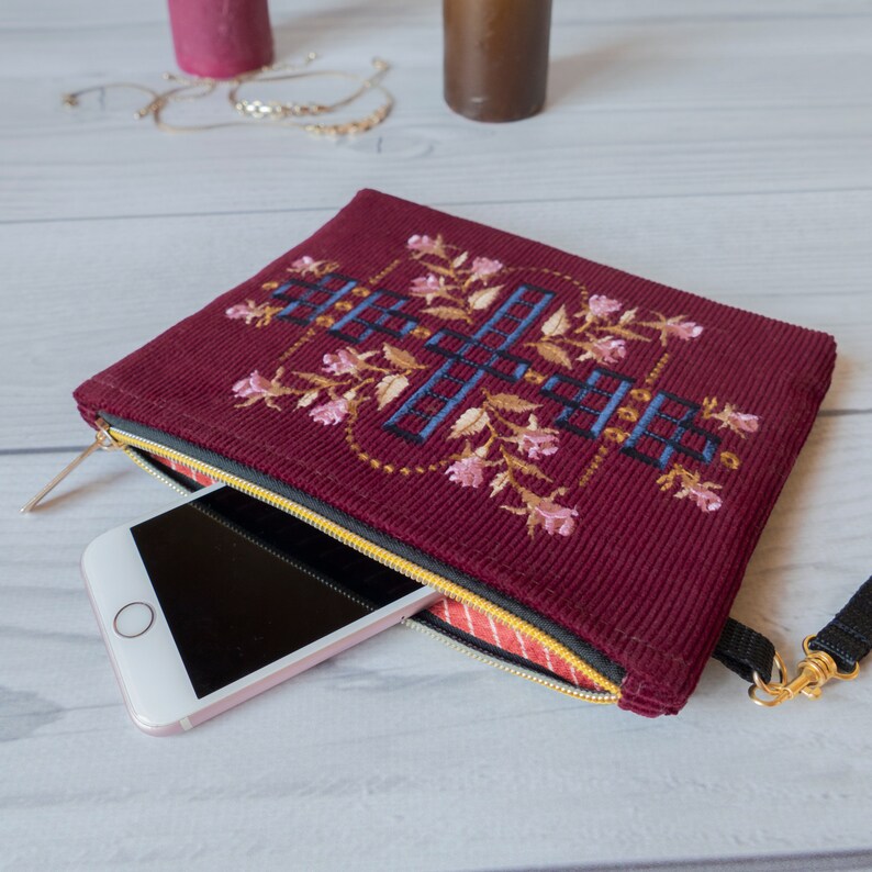 May include: A burgundy corduroy zippered pouch with a floral embroidery design. The pouch has a gold zipper and a black strap. A smartphone is inside the pouch.