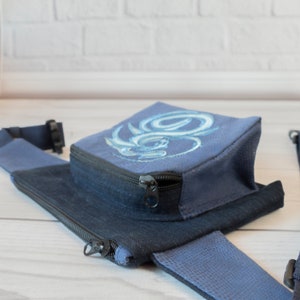 May include: A blue canvas fanny pack with a zipper closure and a blue and white embroidered design. The fanny pack has a black adjustable strap.