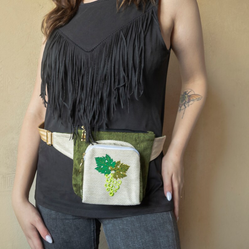 May include: A green and white fanny pack with a zipper closure and a grape design. The fanny pack is worn around the waist of a person wearing a black shirt and jeans.