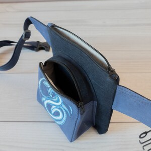 May include: A blue fanny pack with a zippered pocket and a smaller pocket with a white dragon design. The fanny pack has a black strap.