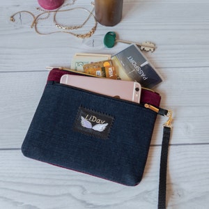 May include: A black and burgundy zippered pouch with a black strap. The pouch has a white embroidered patch with the word "Lidav" and a pair of wings. The pouch is open and contains a smartphone, a credit card, cash, and a passport.