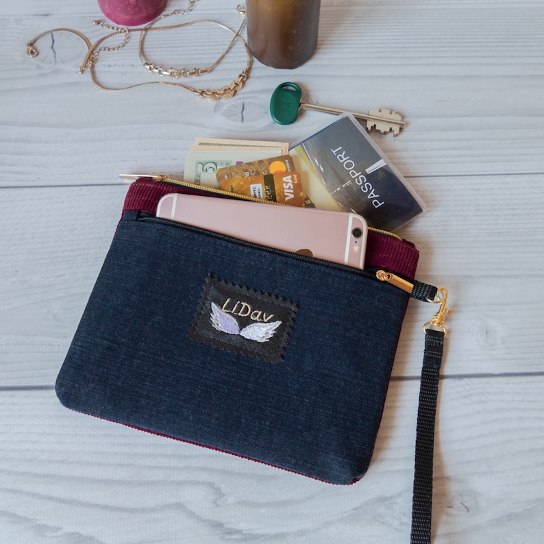 May include: A black and burgundy zippered pouch with a black strap. The pouch has a white embroidered patch with the word "Lidav" and a pair of wings. The pouch is open and contains a smartphone, a credit card, cash, and a passport.