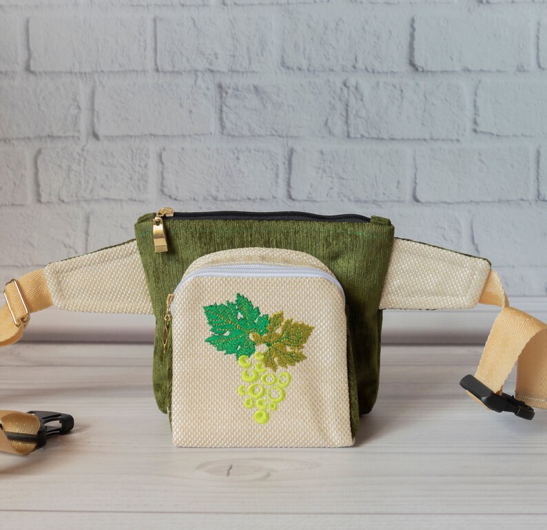 May include: A green and beige fanny pack with a zippered pouch featuring an embroidered grape design. The pouch is attached to the fanny pack with a zipper.