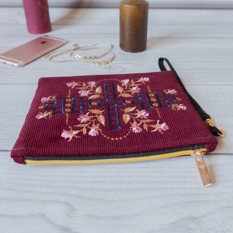 May include: A burgundy corduroy zippered pouch with a black zipper and gold zipper pull. The pouch features an embroidered design of a floral pattern with a geometric centre.