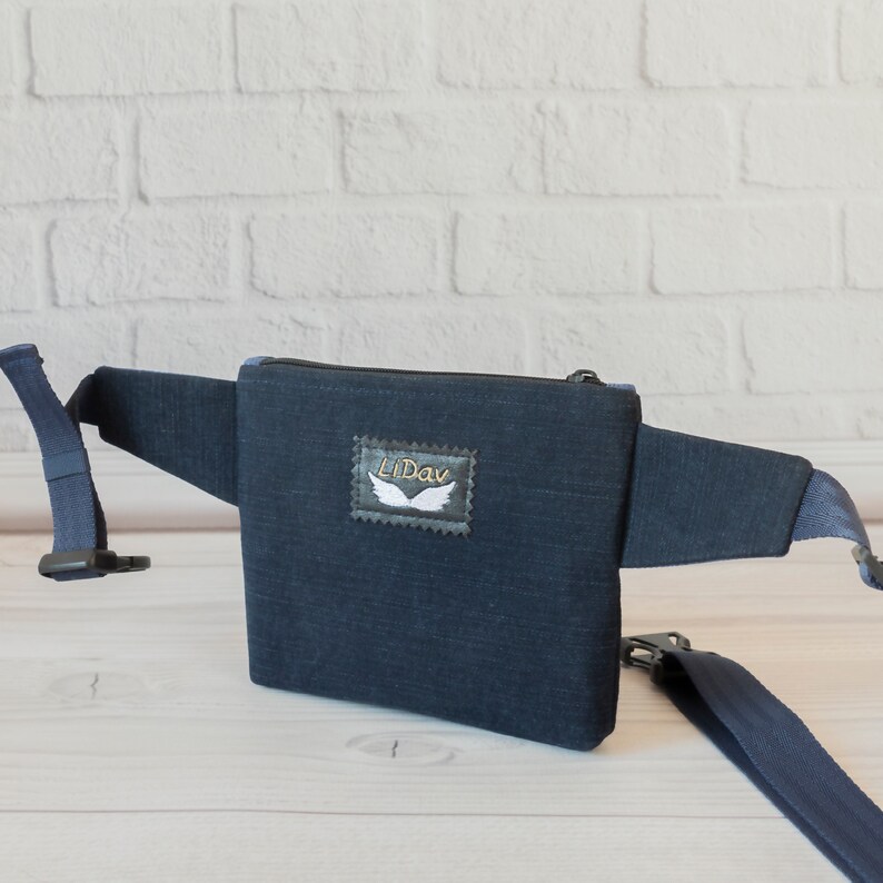 May include: A dark blue denim fanny pack with a zipper closure and a white embroidered wing logo that says "Lidav".