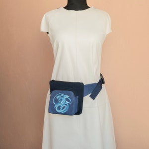 May include: A blue fanny pack with a blue dragon design. The fanny pack is worn around the waist of a person wearing a white dress.