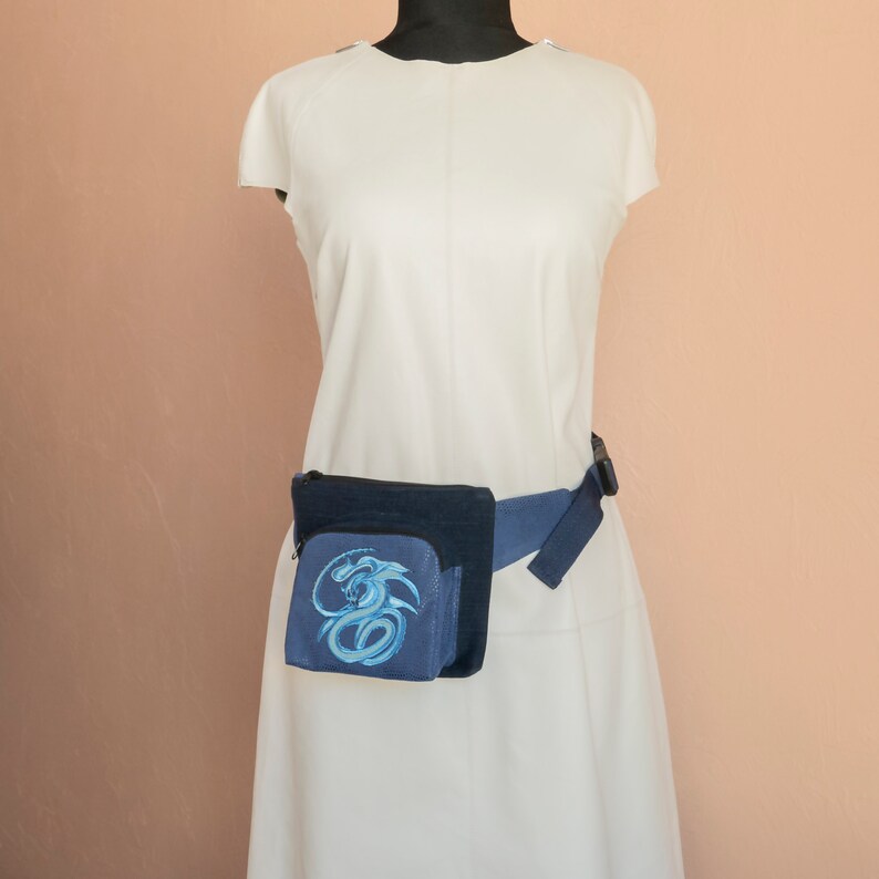 May include: A blue fanny pack with a blue dragon design. The fanny pack is worn around the waist of a person wearing a white dress.