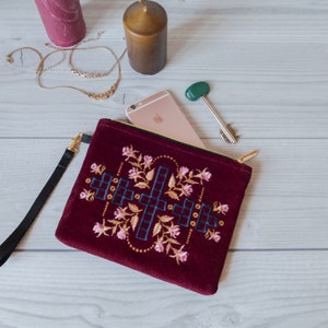 May include: A burgundy velvet clutch with a gold zip and embroidered floral design. The clutch has a geometric pattern in navy blue and white.