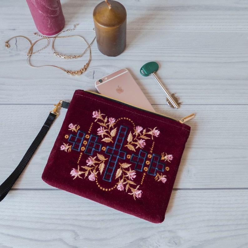 May include: A burgundy velvet clutch with a gold zip and embroidered floral design. The clutch has a geometric pattern in navy blue and white.
