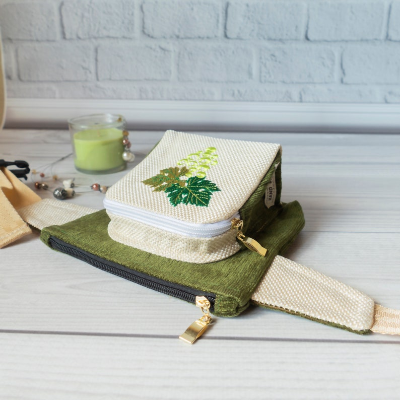 May include: A green and white fabric pouch with a zipper closure and a gold zipper pull. The pouch has a white embroidered design of a grapevine with green leaves. The pouch is attached to a white fabric strap.