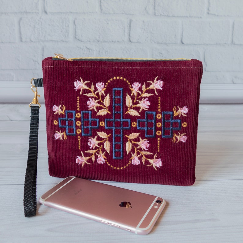 May include: A burgundy corduroy clutch with a gold zip and a black strap. The clutch features an embroidered design of a cross with floral accents in pink and yellow.