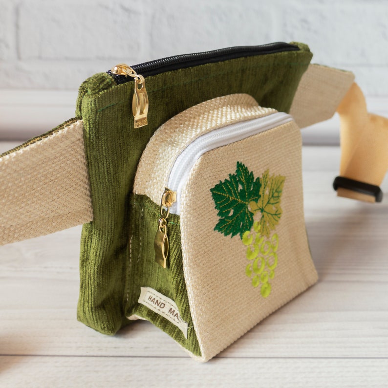 May include: A green and beige fanny pack with a zipper closure and a white zipper pull. The front pocket features an embroidered design of a bunch of grapes with green leaves. The bag has a brown leather strap and the text "HAND MADE" is visible on the bottom.