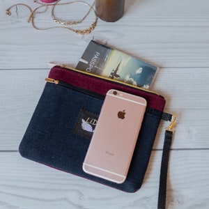 May include: A dark blue and burgundy zippered pouch with a black strap. The pouch has a small embroidered patch with the word "LID" and a wing design. A rose gold iPhone is inside the pouch, along with a small booklet with the word "PASSPO" visible.