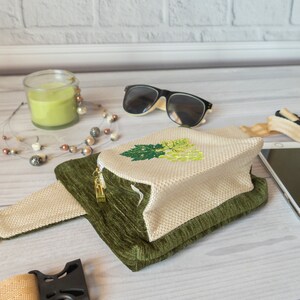 May include: A small, green and beige fanny pack with a zipper closure. The fanny pack has a white embroidered design of a bunch of grapes with green leaves.