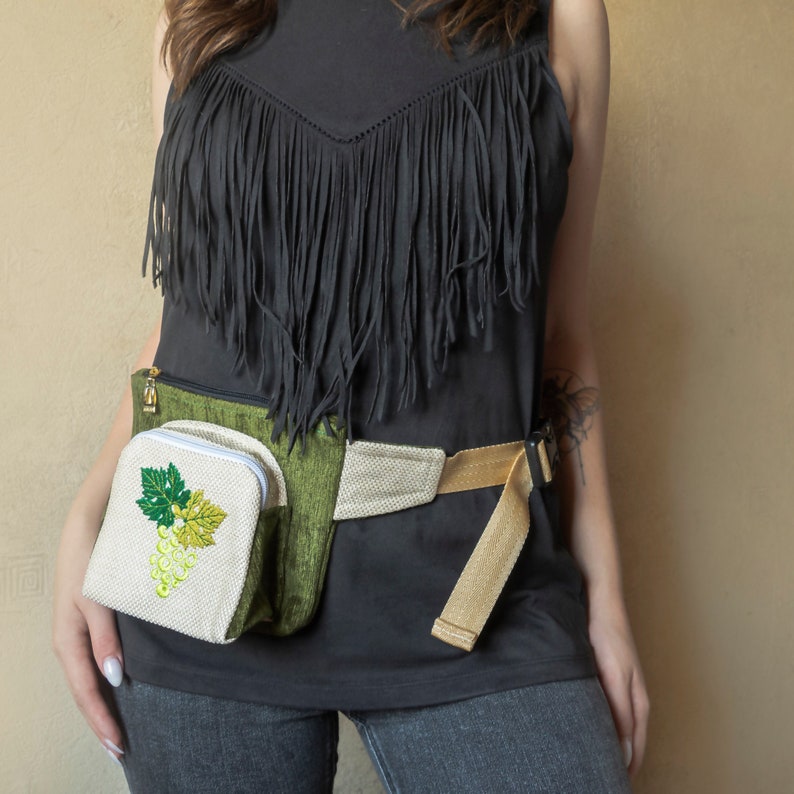 May include: A black fringed top with a green and beige fanny pack featuring an embroidered white grape design.