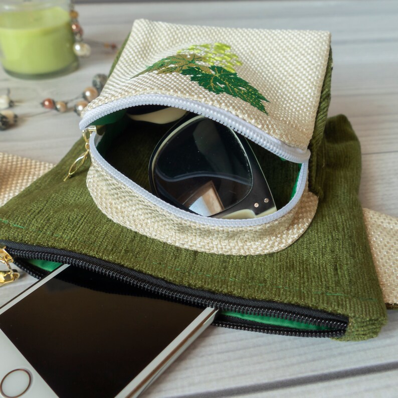 May include: A green and beige zippered pouch with a white zipper pull. The pouch is open and contains a pair of sunglasses and a smartphone. The pouch has a floral embroidery design on the front.