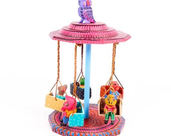 May include: A colourful wooden carousel with four swings, each holding a painted animal figure. The carousel is topped with a purple owl with pink and black details. The base is decorated with intricate patterns in shades of pink, blue, green, and orange.