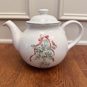 May include: White ceramic teapot with a lid and handle. The teapot features a decorative design of a green plant with a red ribbon and a red bird. The rim of the teapot is trimmed with green.