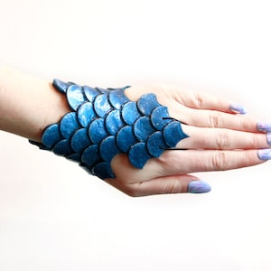 Puede incluir: Un brazalete azul con forma de escamas. El brazalete está hecho de piezas circulares superpuestas que crean una apariencia texturizada. El brazalete se lleva en la muñeca y se extiende parcialmente sobre la mano. Las escamas son de color azul vibrante.