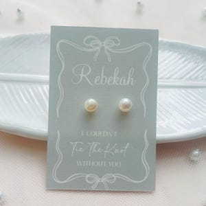 May include: Pearl stud earrings presented on a pale blue card, inscribed with "Rebekah" and the message "I Couldn't Tie The Knot Without You". The card rests on a white decorative plate.