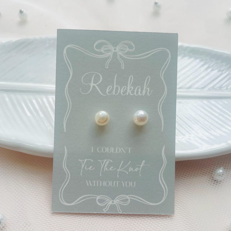 May include: Pearl stud earrings presented on a pale blue card, inscribed with "Rebekah" and the message "I Couldn't Tie The Knot Without You". The card rests on a white decorative plate.