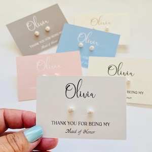 Puede incluir: Una mano sostiene una tarjeta blanca con el nombre "Olivia" en cursiva y dos pendientes de perlas. La tarjeta dice "THANK YOU FOR BEING MY Maid of Honor." Otras tarjetas en varios colores pastel con el nombre "Olivia" y pendientes están en el fondo.
