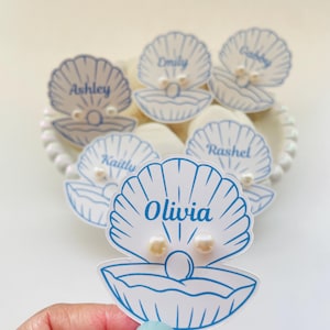 May include: White seashell-shaped place cards with blue outlines and names like "Olivia" and "Ashley" written in blue script. Each card has a pearl accent. The cards are arranged on a white plate.
