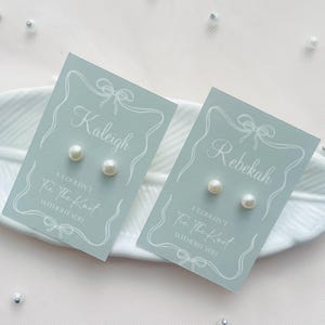 May include: Two pairs of pearl stud earrings on light blue cards. Each card features a name, decorative border, and the text "I couldn't tie the knot without you". The earrings are presented on a white, feather-shaped dish.