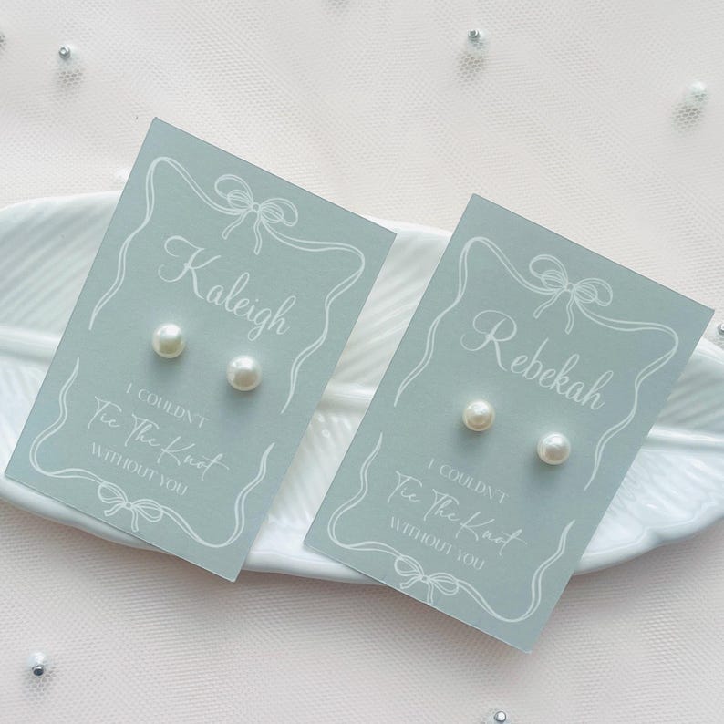 May include: Two pairs of pearl stud earrings on light blue cards. Each card features a name, decorative border, and the text "I couldn't tie the knot without you". The earrings are presented on a white, feather-shaped dish.