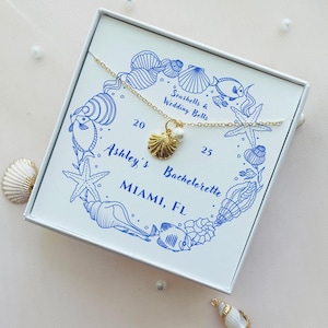 May include: A gold-colored necklace with a seashell pendant and a small pearl, presented in a white box. The box is decorated with blue illustrations of seashells, starfish, and fish, with text that reads "Ashley's Bachelorette MIAMI, FL."