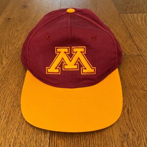 Puede incluir: Gorra de béisbol granate y dorada con un logotipo "M" dorado bordado. La gorra tiene una corona granate y una visera dorada. El botón superior es dorado. Un accesorio deportivo clásico.