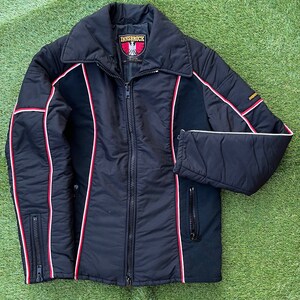 May include: A black quilted jacket with red and white piping. The jacket has a zipper closure and two pockets. The label on the jacket reads "Innsbruck".