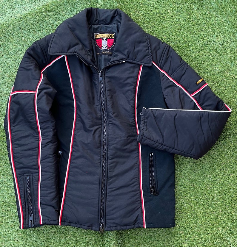 May include: A black quilted jacket with red and white piping. The jacket has a zipper closure and two pockets. The label on the jacket reads "Innsbruck".