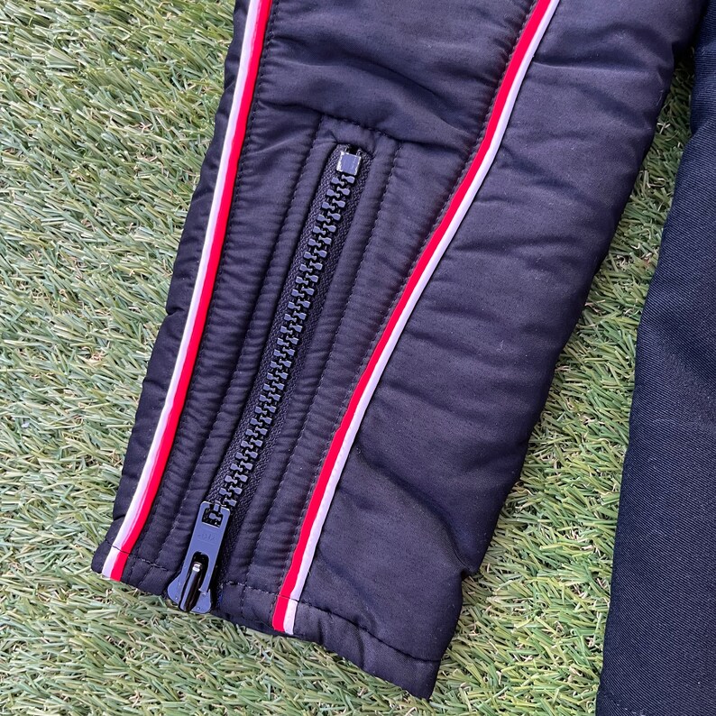 May include: Black puffer jacket with red and white piping detail. The jacket has a zipper closure and a quilted design.