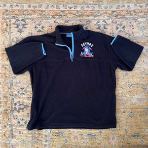 May include: Black fleece pullover with a blue zip and blue trim. The front of the shirt features a cartoon image of Eeyore from Winnie the Pooh with the text "Eeyore Feeling Gloomy Since 1948".
