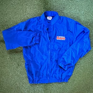 May include: A blue windbreaker jacket with a zipper closure and elastic cuffs. The jacket has the logo "MYRINA TURBO DIESEL ENGINE OIL" embroidered on the front.