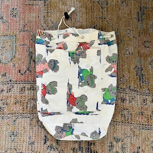 Puede incluir: Bolsa blanca con cordón con un patrón repetido de koalas grises con trajes coloridos. Los koalas se representan en varias poses. La bolsa tiene un pequeño bolsillo y un cierre de cordón.