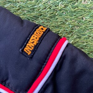 May include: Close-up of a black jacket with a red and white trim. The jacket has a yellow embroidered patch that says "Dunsford".