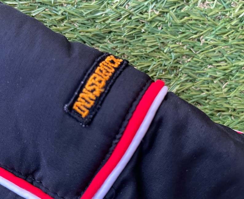 May include: Close-up of a black jacket with a red and white trim. The jacket has a yellow embroidered patch that says "Dunsford".