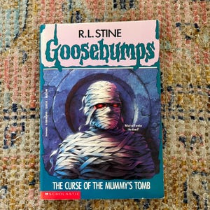 Puede incluir: Un libro de bolsillo titulado "Goosebumps: The Curse of the Mummy's Tomb" de R.L. Stine. La portada presenta una momia con ojos rojos brillantes. El libro tiene un esquema de color turquesa y rosa con el texto "What will wake the dead?"