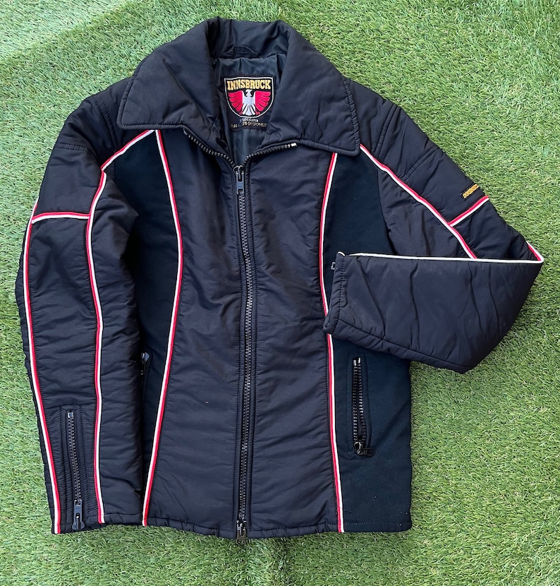 May include: A black ski jacket with red and white piping. The jacket has a zipper closure and two pockets. The label on the jacket reads "Innsbruck".
