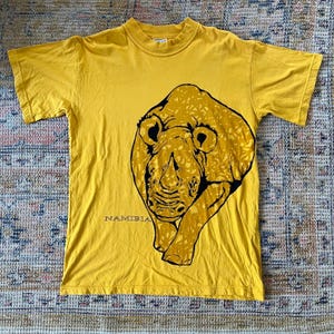 Peut inclure: Un t-shirt jaune avec un imprimé de rhinocéros noir et jaune et le mot "NAMIBIA" imprimé sur le devant.