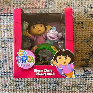 Puede incluir: Un reloj despertador y una alcancía de Dora la Exploradora de color rosa y verde con Dora y Botas. La esfera del reloj tiene una imagen de una escena de la selva con las palabras "Camino" y "Dora" en ella. El embalaje es rosa con las palabras "Nick Jr. Dora la Exploradora" en él.
