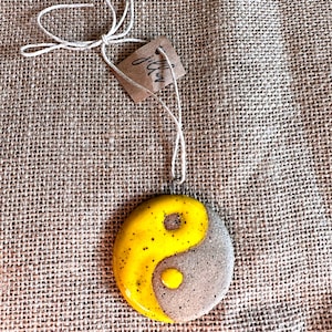Könnte beinhalten: Ein gelber und grauer Keramik-Yin-Yang-Symbol-Anhänger mit einer Schnur. Der Anhänger liegt auf einem braunen Jutesack-Hintergrund.