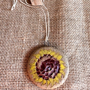 Op de afbeelding: Een rond keramiek ornament met een geschilderd zonnebloemontwerp. De zonnebloem heeft een bruin centrum en gele bloemblaadjes. Het ornament hangt aan een wit touwtje.