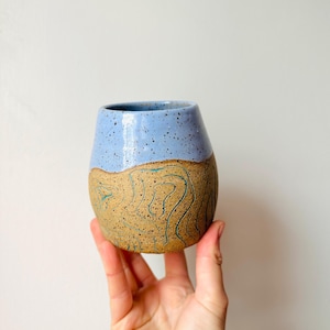 Könnte beinhalten: Eine hellblaue und braune Keramiktasse mit einem gesprenkelten Überzug und einem wellenförmigen Design.